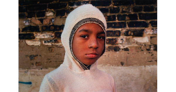 A color portrait photograph of a young dark-skinned child wearing a white hooded sweater with the hood pulled tight around their head.
