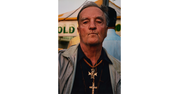 A color portrait photograph of a medium-skinned man with gray hair, lips pursed, wearing several gold cross necklaces over a black shirt.