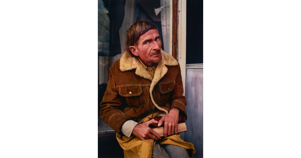 A color portrait photograph of a seated middle-aged White man, short hair, a stubbly goatee, wearing a brown coat lined with cream fleece, holding a book in his lap.