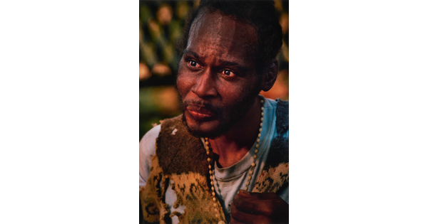 A color portrait photograph of a Black man with a goatee wearing a beaded necklace, and a torn cutoff vest over a t-shirt.