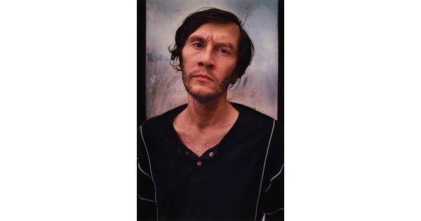 A color portrait photograph of a White man with short untidy hair, a stubbly beard and mustache, a furrowed brow, wearing a black open-necked shirt.