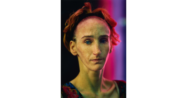 A color portrait photograph of a White woman with a headband pulling back her short auburn hair, a freckled face, and prominent cheekbones.
