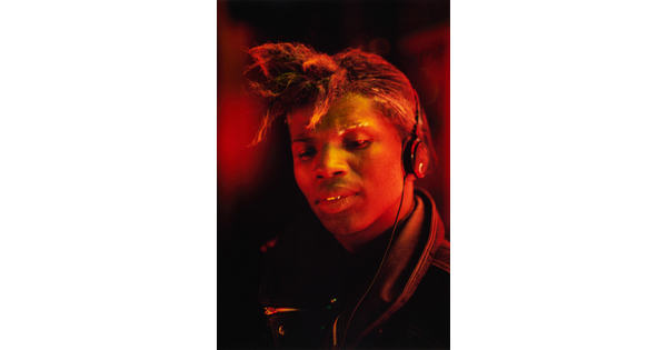 A color portrait photograph of a Black man, hair pulled into a sort of ponytail over his forehead, wearing headphones, bathed in reddish-orange light.