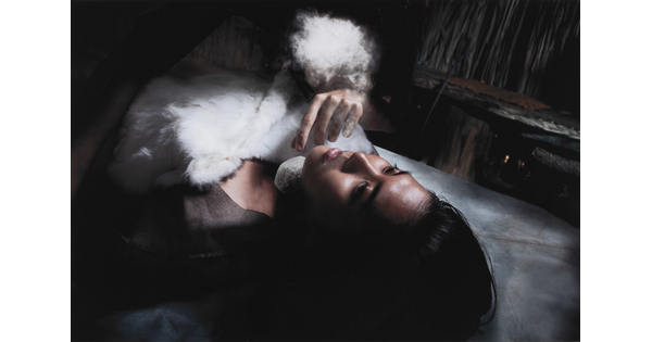 A color photograph of a young Indigenous woman lying on her back, eyes open, chest obscured by fluffy cloudlike shapes, as a pale hand reaches toward her face.