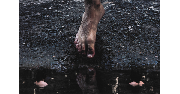 A color photograph of a masculine foot lifting off a muddy, gravelly bank about to step into the dark water in which it is reflected.