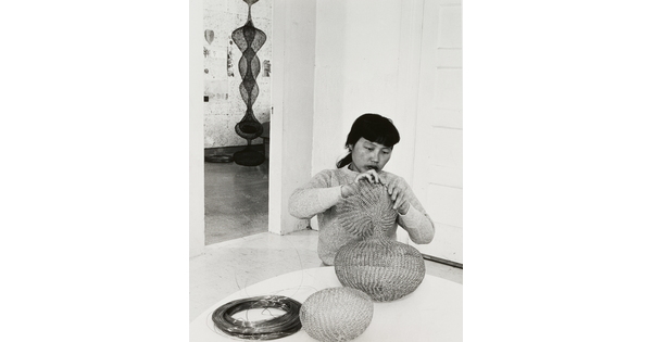 A black-and-white photograph of a woman with Asian features seated at a table and working an a wire sculpture.