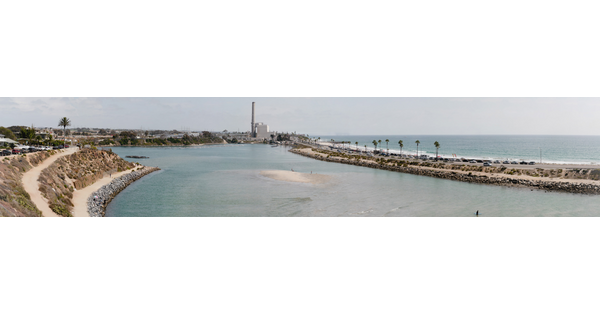A panoramic color photograph of (from left to right) a rocky shoreline, a shallow bay, a narrow isthmus with traffic leading to a power plant, and a larger body of water.