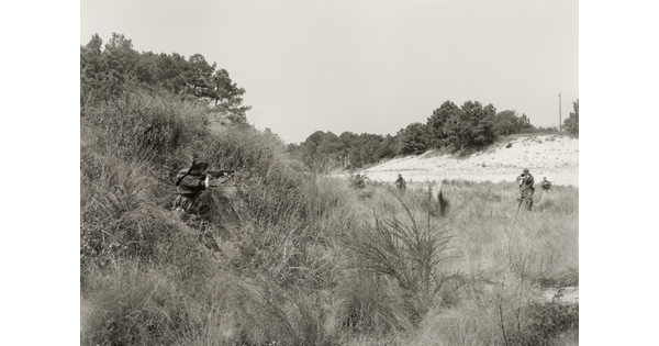 A black-and-white photograph of a person in military clothing crouching in tall grass and aiming a rifle at several other soldiers in a nearby field.