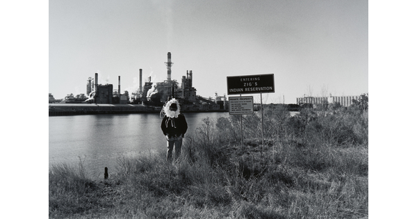A black-and-white photograph of a man, face in shadow, wearing a feathered headdress standing in grass on the banks of a body of water next to a sign that reads, "Zig's Indian Reservation"; across the water is an industrial facility.