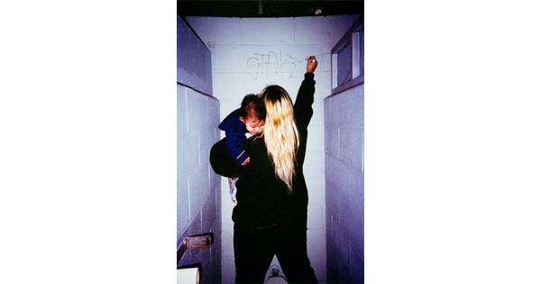 A color photograph of a woman, her back to the viewer, holding a baby while writing graffiti on a bathroom stall wall.