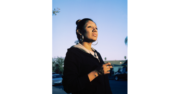 A color photograph of a medium-skinned woman, hair pulled on top of her head and large hoop earrings, standing outside holding a cigarette in her hand.