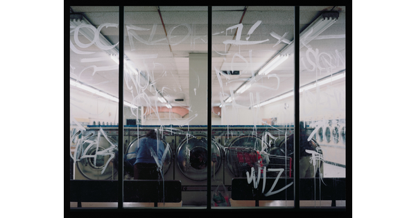 A color photograph of a laundromat at night through as seen through glass windows covered in white graffiti.