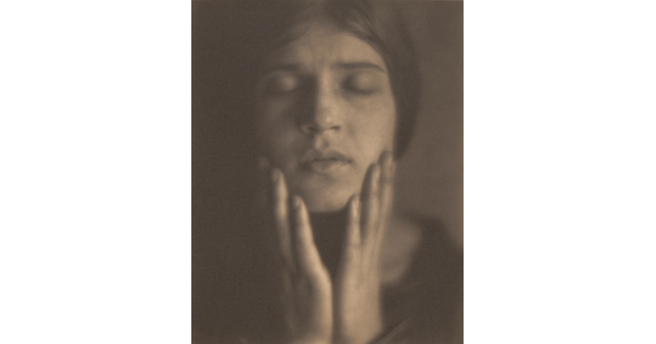 A black-and-white photograph of the face of a fair-skinned woman, eyes closed, hair pulled back, holding her hands to her face.