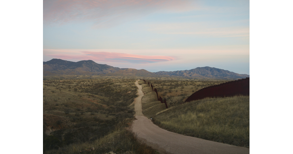 A color photograph of a dirt road that follows along a tall wall through rolling hills with mountains in the distance.