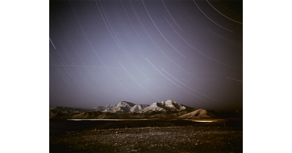 A color photograph of a dark desert landscape and mountains against a dark blue night sky with light streaks curving across the sky.