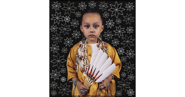 A color photograph of a young Indigenous girl, dark hair pulled back, wearing a yellow shirt, beaded necklaces, turquoise jewelry, holding a feathered fan, standing in front of a black background with white stars.