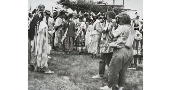 A black-and-white photograph of two women photographing a group of Indigenous people in cultural dress.