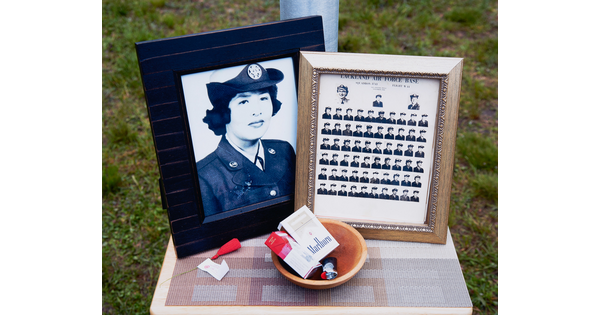 A color photograph of a table with two framed black-and-white photos: one of an Indigenous woman in military uniform, the other a class photo, and a wood bowl containing two packs of cigarettes and a lighter.