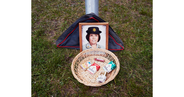 A color photograph of a framed color photo of an Indigenous woman in military uniform behind a basket containing four packs of cigarettes and a lighter placed on grass.