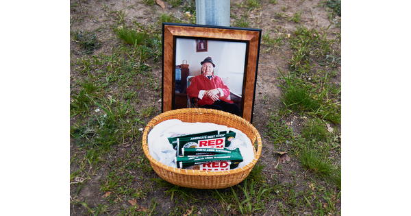A color photograph of a framed color photo of a seated man in a red jacket behind a basket on grass filled with packets of chewing tobacco.