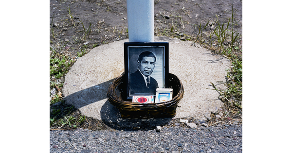 A color photograph of a woven basket on concrete containing a black-and-white framed photo of a man in a suit and two packs of cigarettes at the base of a pole.