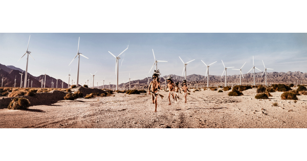 A color photograph of a group of Indigenous children in traditional dress running through a desert landscape covered with wind turbines.