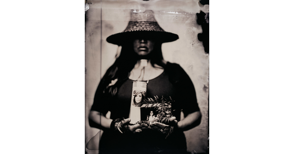 A black-and-white photograph of an Indigenous woman, eyes obscured by a straw hat, holding evergreen leaves, a candle, and photographs in her arms.