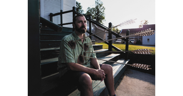 A color photograph of a White man sitting casually on wood steps with large arrows sticking out of his chest.