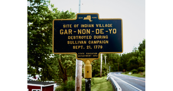 A color photograph of a New York historical marker that reads, "Site of Indian Village Gar-Non-De-Yo Destroyed during Sullivan Campaign Sept. 21, 1779," next to a road.