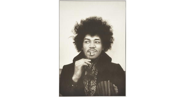 A black-and-white portrait photograph of a Black man with an Afro, looking to the side, holding a pair of sunglasses with the temple tip between his lips.