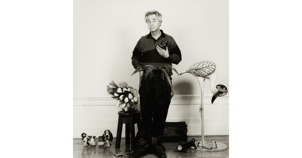 A black-and-white photograph of a White man standing with a camera in one hand, surrounded by plants and whimsical animal sculptures.