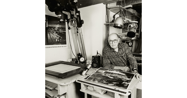 A black-and-white photograph of an older White man looking at the viewer from a photography studio work space.