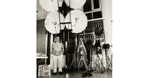 A black-and-white photograph of a balding White man standing with hands in pockets among large lighting umbrellas and other equipment.