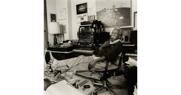 A black-and-white photograph of an older Black man with gray hair and a mustache holding a pipe to his lips, reclining in a chair in a cluttered office.