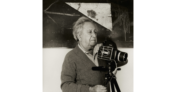 A black-and-white photograph of an older White man holding a camera on a tripod in front of a white and black wall.
