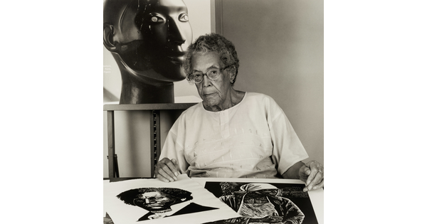 A black-and-white photograph of an older Black woman with two prints on a table in front of her and a large black sculpture of a head behind her.