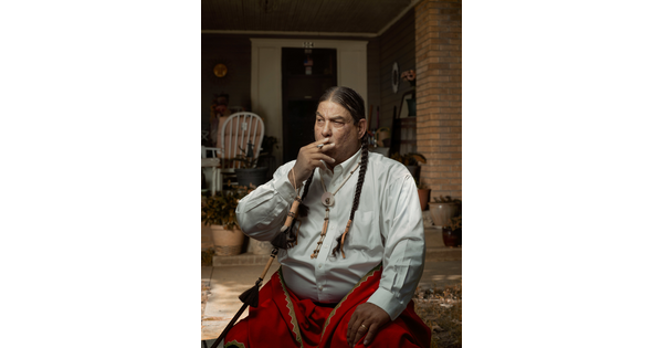 A color photograph of a seated Native American man with two, long braids smoking a cigarette in front of a porch.
