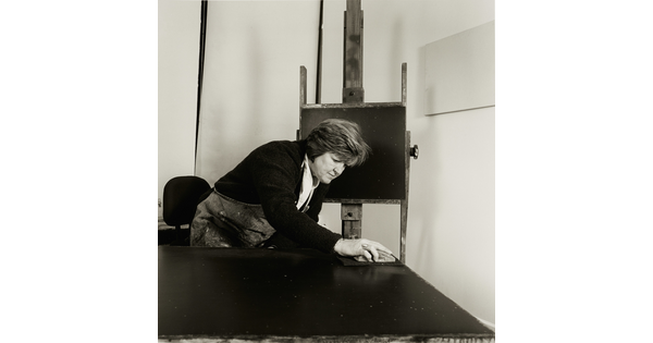 A black-and-white photograph of a White woman working on a large painting with an easel displaying a small painting behind her.