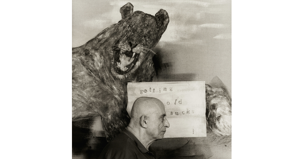 A black-and-white photograph of an older White man in profile standing in front of a painting of a lion holding a sign that says, "getting old sucks."