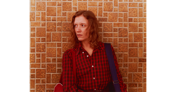 A color photograph of a White woman with reddish hair wearing a plaid, button-down shirt standing against a tile wall and looking to her right.