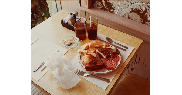 A color photograph of a dining table set with a full plate of food on a white paper placemat, full drinking cups, and an ashtray.