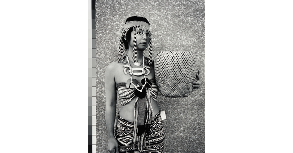 A black-and-white photograph of a medium-skinned woman wearing a headband with long beaded fringes, a halter top, patterned skirt, and holding a woven basket.