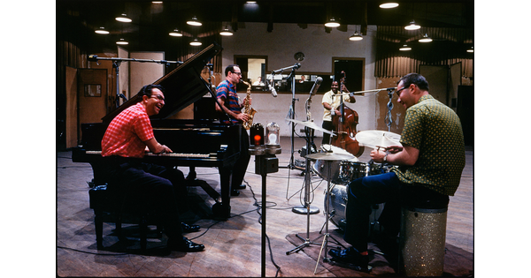 A color photograph of four men, three White and one Black, in a recording studio: one playing a piano, one a saxophone, one the drums, and one playing an upright bass.