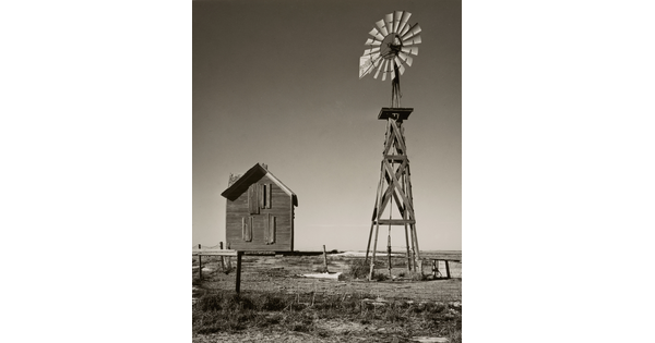 A black-and-white photograph of an abandoned boarded up farmhouse and a windmill.