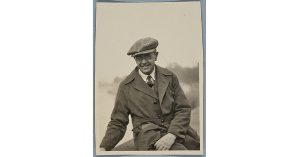 A black-and-white photograph of a smiling White man seated outside, wearing a newsboy cap, round wire-rimmed glasses, and a trench coat.