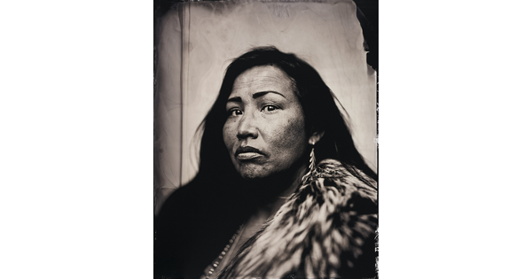 A black-and-white portrait photograph of a Native American woman wearing a fur and long traditional earrings.