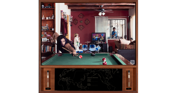 A color photograph of a medium-skinned woman playing pool; in the background, a young child stands next to a blue drum set watching TV, and a man looks through a window from the outside.