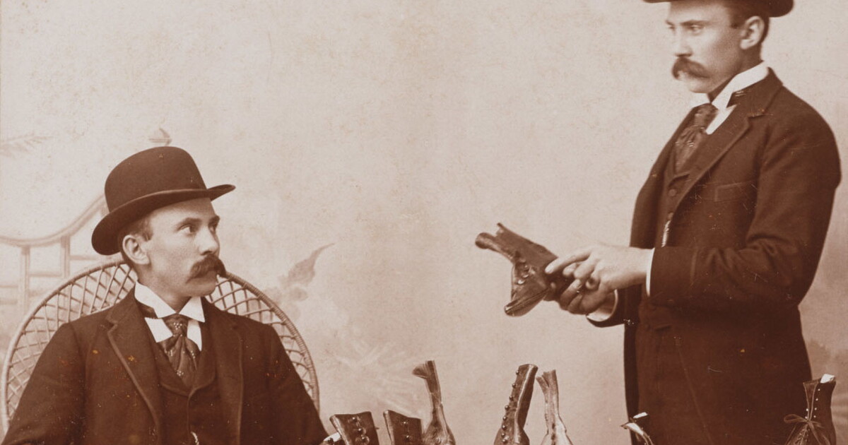 A black-and-white studio photograph of a seated White man in a bowler hat looking at shoes as the same man standing appears to show him shoes.