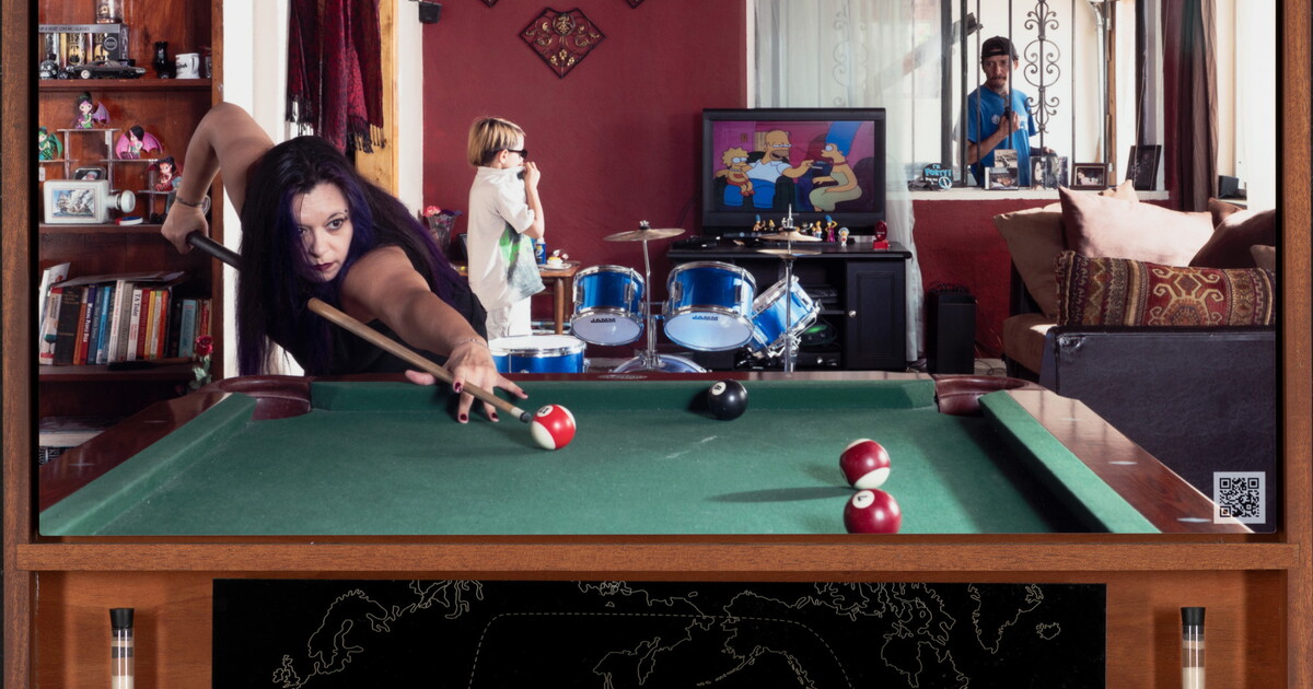 A color photograph of a medium-skinned woman playing pool; in the background, a young child stands next to a blue drum set watching TV, and a man looks through a window from the outside.