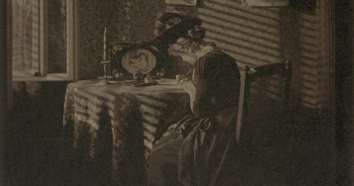 A sepia-toned photograph of a seated woman writing at a table, illuminated by light from a window.